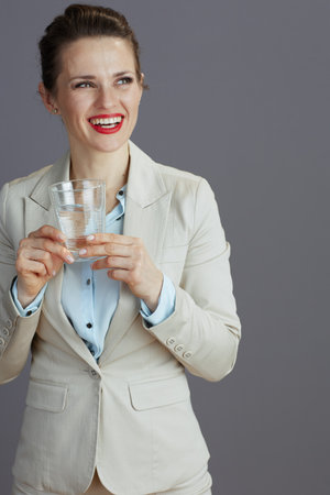 happy modern business woman in a light business suit with glass of water isolated on gray.の写真素材