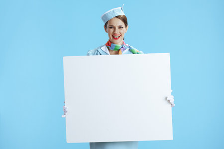 happy elegant air hostess woman against blue background in blue uniform showing blank board.の写真素材