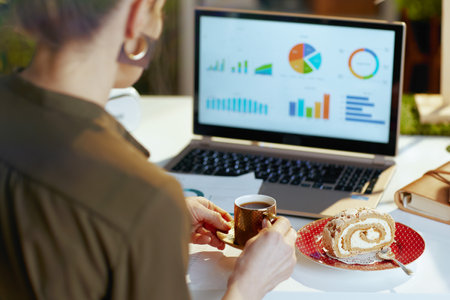 Seen from behind modern female with documents, laptop, piece of cake and cup of coffee in modern office.の写真素材
