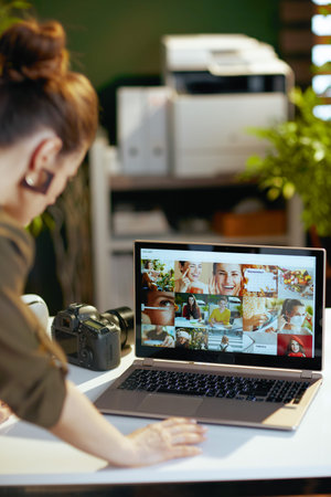 Seen from behind female photographer with laptop and photo camera viewing photos in modern office.