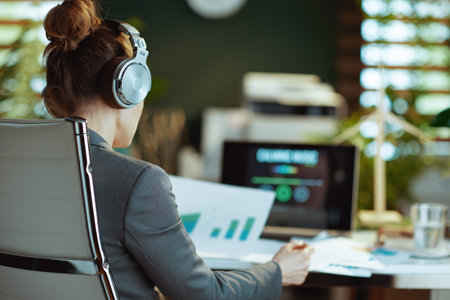 Seen from behind modern woman with documents listening to music with headphones in modern office.の写真素材