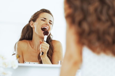 Happy young woman singing in makeup brush in bathroomの写真素材