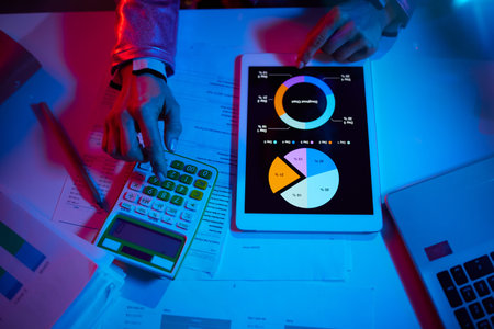 Upper view of modern woman with calculator and documents exploring graphs and using tablet PC in...