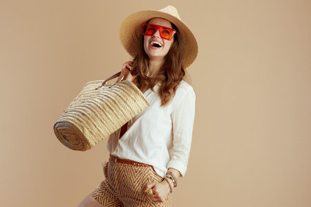 Beach vacation. happy trendy woman in white blouse and shorts isolated on beige with straw bag, summer hat and sunglasses.の写真素材