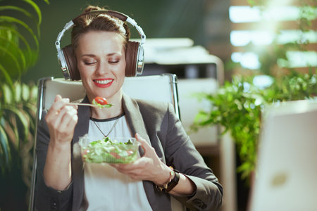 Sustainable workplace. smiling modern 40 years old bookkeeper woman in a gray business suit in modern green office with laptop eating salad and listening to the music with headphones.の写真素材