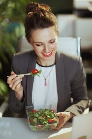 Sustainable workplace. smiling modern middle aged accountant woman in a gray business suit in modern green office eating salad.の写真素材