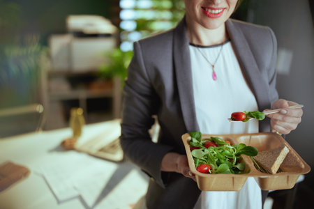 Sustainable workplace. happy modern woman worker in a gray business suit in modern green office with laptop eating salad.の写真素材