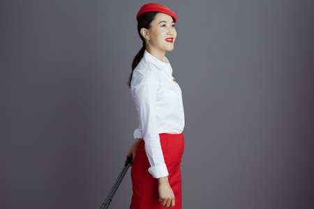 smiling elegant asian female flight attendant in red skirt and hat uniform with trolley bag looking into the distance isolated on gray background.の写真素材