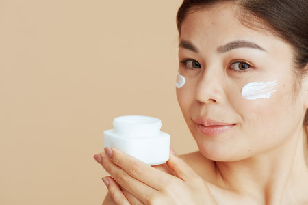 modern woman with facial cream jar and facial cream on face isolated on beige background.の写真素材