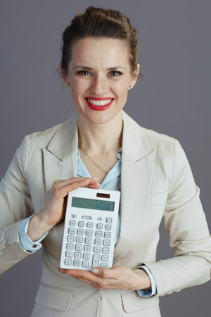 smiling modern middle aged woman worker in a light business suit with calculator against gray background.の写真素材