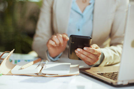 Closeup on small business owner woman in a light business suit in green office with laptop using smartphone.の写真素材