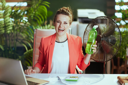 Sustainable workplace. Portrait of happy modern 40 years old accountant woman at work in red jacket with bottle of water, electric fan and laptop.の写真素材