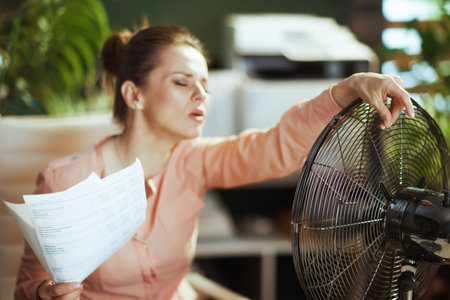 Sustainable workplace. modern female employee in modern green office with documents and electric fan suffering from summer heat.の写真素材
