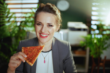 Sustainable workplace. smiling modern female employee in a gray business suit in modern green office with pizza.の写真素材