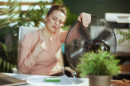 Sustainable workplace. modern 40 years old woman worker in modern green office with electric fan...