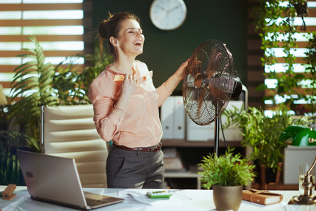 Sustainable workplace. happy modern 40 years old accountant woman in modern green office with electric fan and laptop.の写真素材