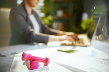 Sustainable workplace. Closeup on modern small business owner woman in modern green office with dumbbells and laptop.の写真素材