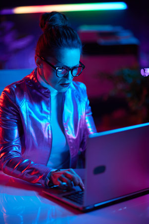 Neon metaverse futuristic concept. elegant middle aged woman in glasses using laptop in modern office.の写真素材
