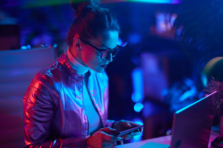 Neon metaverse futuristic concept. elegant female in glasses with laptop using smartphone in modern office.の写真素材