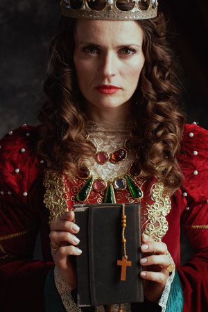 medieval queen in red dress with book, rosary and crown on dark gray background.の写真素材
