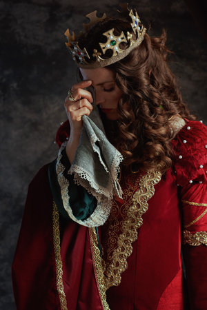 medieval queen in red dress with handkerchief and crown crying on dark gray background.の写真素材