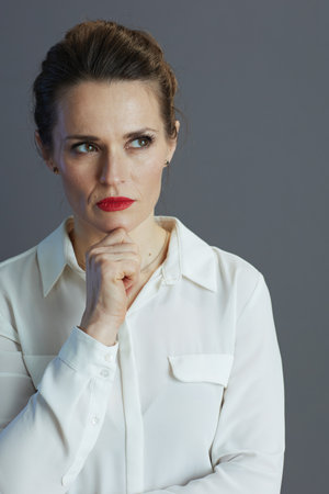 pensive elegant 40 years old woman worker in white blouse looking aside against gray background.の写真素材