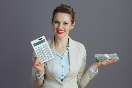 happy stylish middle aged woman worker in a light business suit with calculator and dollars money packs isolated on grey.の写真素材