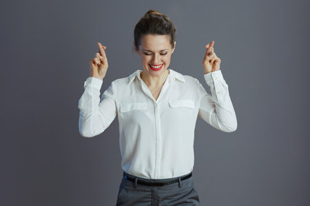 happy stylish female employee in white blouse with crossed fingers against gray background.の写真素材