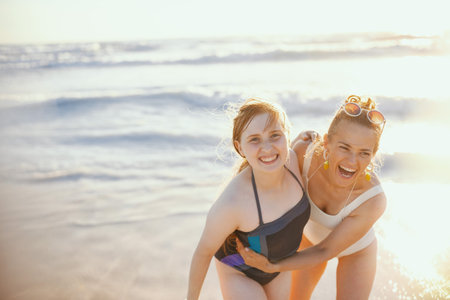 Portrait of happy modern mother and teenage daughter at the beach in swimsuit having fun time.