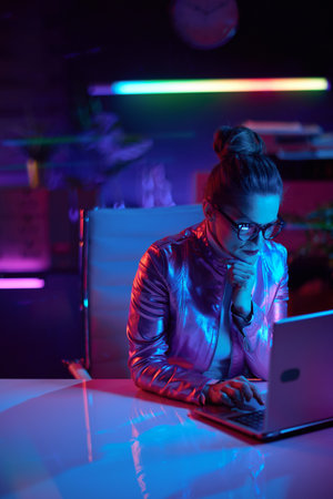 Neon metaverse futuristic concept. pensive elegant business woman in glasses using laptop in modern office.の写真素材