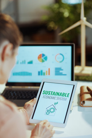 Seen from behind modern woman with laptop exploring charts and using digital tablet in modern office.の写真素材