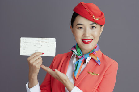 happy stylish air hostess asian woman in red skirt, jacket and hat uniform with flight tickets isolated on gray background.の写真素材