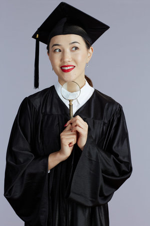 pensive young graduate student asian woman with magnifying glass isolated on gray background.