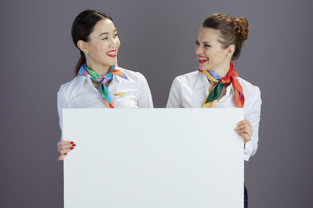 smiling elegant air hostess women in blue skirt, white shirt and scarf with blank billboard against gray background.の写真素材