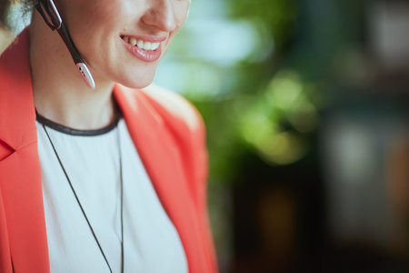 Sustainable workplace. Closeup on happy modern business woman in modern green office in a red jacket with headset.の写真素材