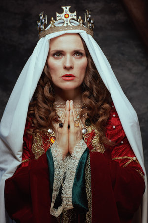medieval queen in red dress with veil and crown praying on dark gray background.の写真素材