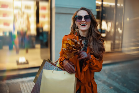 Hello September. smiling middle aged woman in brown trench coat with shopping bags and autumn yellow leaves near shop in the city.の写真素材