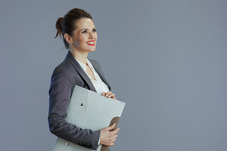 smiling trendy 40 years old woman worker in gray suit with folder isolated on gray background.の写真素材