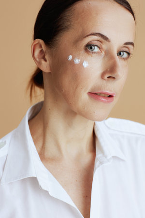 modern middle aged woman with eye cream on face in white shirt on beige background.の写真素材