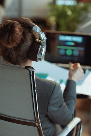 Seen from behind modern business woman with laptop listening to music with headphones in modern office.の写真素材
