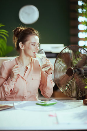 Sustainable workplace. happy modern 40 years old woman employee at work with electric fan, cup of water and lemon.の写真素材