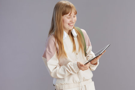 happy trendy teenager girl in beige tracksuit with headphones using tablet PC isolated on grey.
