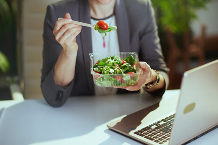Sustainable workplace. Closeup on woman worker in a gray business suit in green office with laptop eating salad.の写真素材