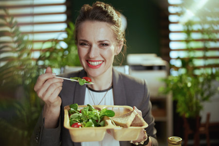 Sustainable workplace. smiling modern middle aged woman employee in a gray business suit in modern green office eating salad.の写真素材