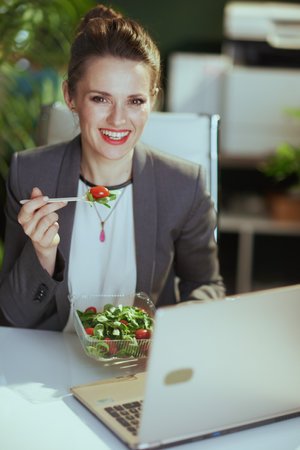 Sustainable workplace. smiling modern female employee in a gray business suit in modern green office with laptop eating salad.の写真素材