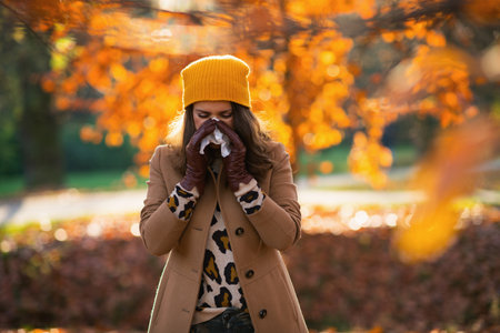 Hello September. elegant middle aged woman in beige coat and orange hat with napkin blowing nose outside on the city park in autumn.の写真素材