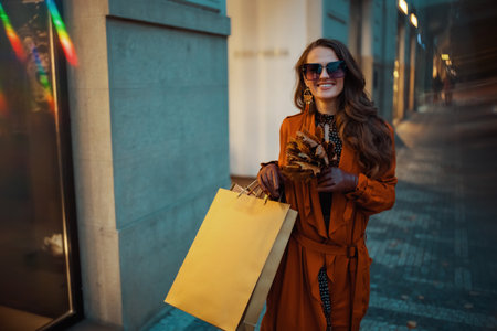 Hello September. happy trendy female in brown trench coat with shopping bags and autumn yellow leaves near store in the city.の写真素材