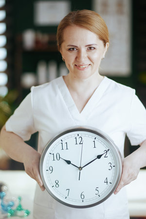 Healthcare time. happy massage therapist woman in massage cabinet with clock.の写真素材