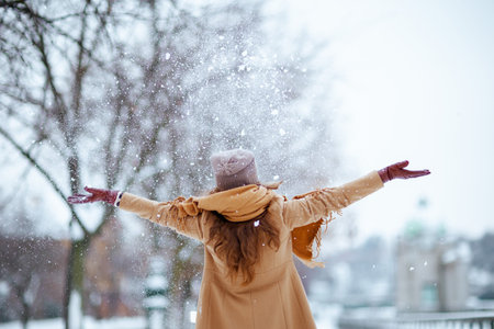 Seen from behind middle aged woman in brown hat and scarf in camel coat with gloves catching snow outside in the city in winter.の写真素材