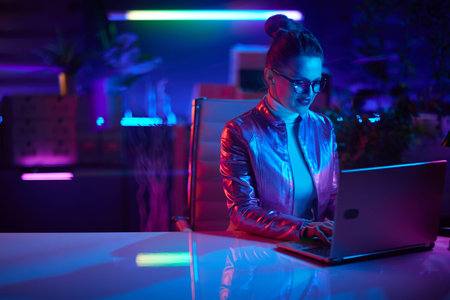 Neon metaverse futuristic concept. happy trendy business woman in glasses using laptop and having video call in modern office.の写真素材